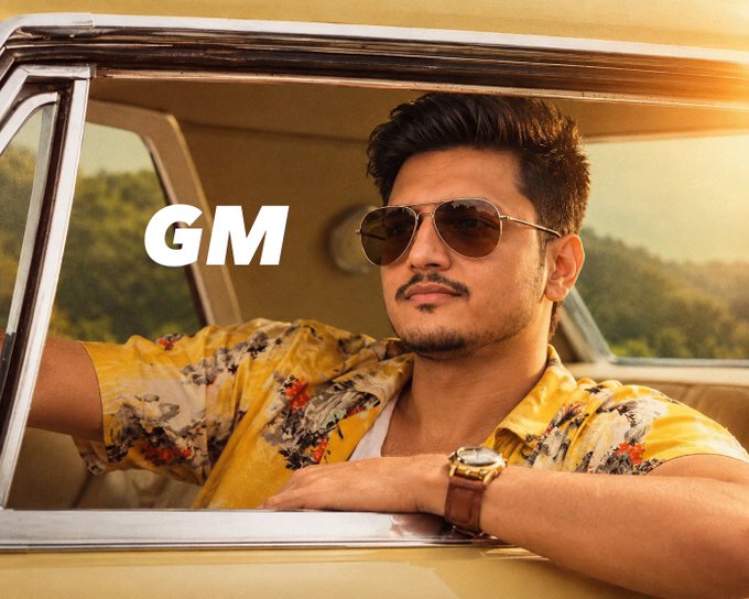 A man wearing aviator sunglasses, a yellow floral Hawaiian shirt, and a gold watch leans out of a car window with the text 'GM' overlaid on the image.