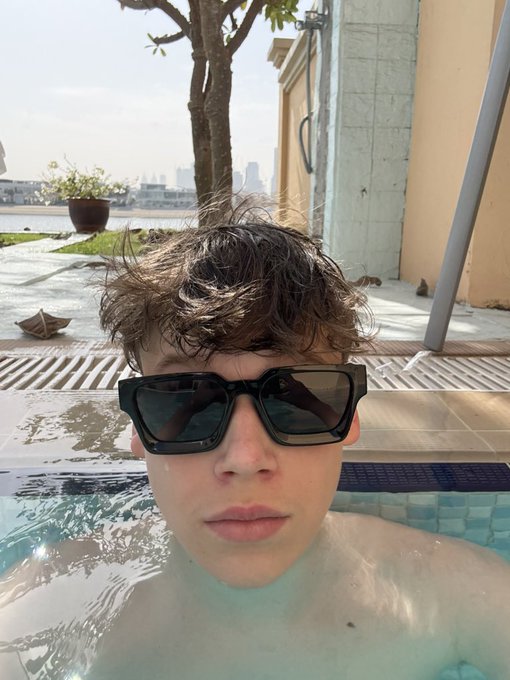 A selfie of a young person with curly brown hair wearing black sunglasses while partially submerged in a swimming pool, with a waterfront cityscape in the background.