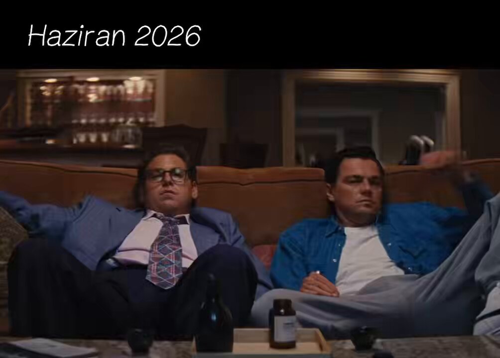 The image shows two men sitting on a couch, one wearing glasses and the other wearing a blue shirt. They are both holding drinks, with wine glasses visible near them. The background includes a dining table and chairs, suggesting that they might be at home or in a casual setting.