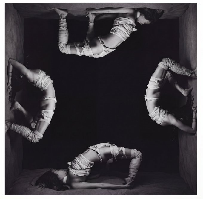 Four contorted figures wrapped in white cloth are arranged along the edges of a dark, box-like space, their arched bodies facing inward to form a central void. The symmetrical, bound poses visually echo the post's Dhammapada quote about samsara, evoking entrapment and the cyclical nature of desire as the figures appear caught and suspended within the frame.