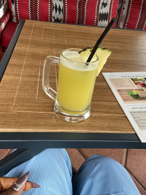 A refreshing pineapple mocktail garnished with a pineapple wedge and black straw sits on a wooden table beside a "JUICES & MOCKTAILS" menu, while a person in a striped shirt relaxes with manicured nails visible—capturing a laid-back "touch grass" moment to kick off the new month.