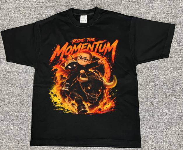 A black t-shirt laid on a gray surface featuring a graphic design with the text 'RIDE THE MOMENTUM' depicting a character with glasses riding a fiery bull.