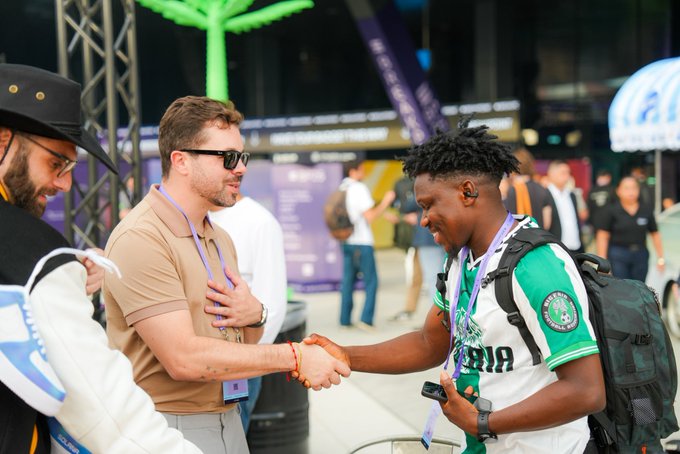 Two men shake hands warmly at what appears to be a crypto/blockchain conference, with one wearing a green and white "INA" jersey and a lanyard, while the other sports a tan polo and sunglasses. The image captures the meaningful personal connection referenced in the post "GM Solana - Crazy how your life can change by just meeting people," illustrating how a simple handshake at an event can spark relationships that transform lives.