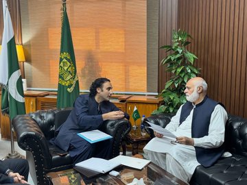 Two men sit in leather chairs in an office setting with Pakistani flags, engaged in a discussion over documents.