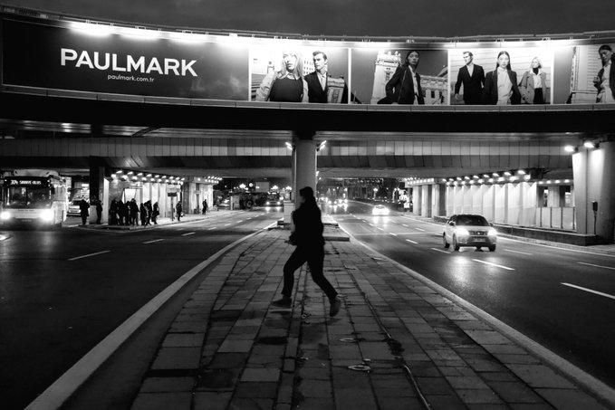 The image shows a person walking down a sidewalk at night, with a large billboard featuring several people in suits above them. The scene takes place under an overpass or bridge, which can be seen in the background of the photo. A car is also visible on the street below the billboard. The overall atmosphere suggests that it might be nighttime and the person walking down the sidewalk could be heading to a nearby location such as work or another destination.