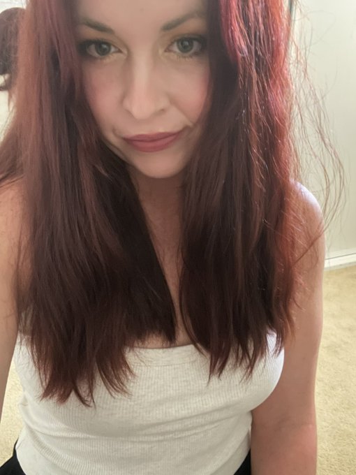 A selfie of a woman with long auburn/red hair wearing a white tank top, looking at the camera with a slight smile.