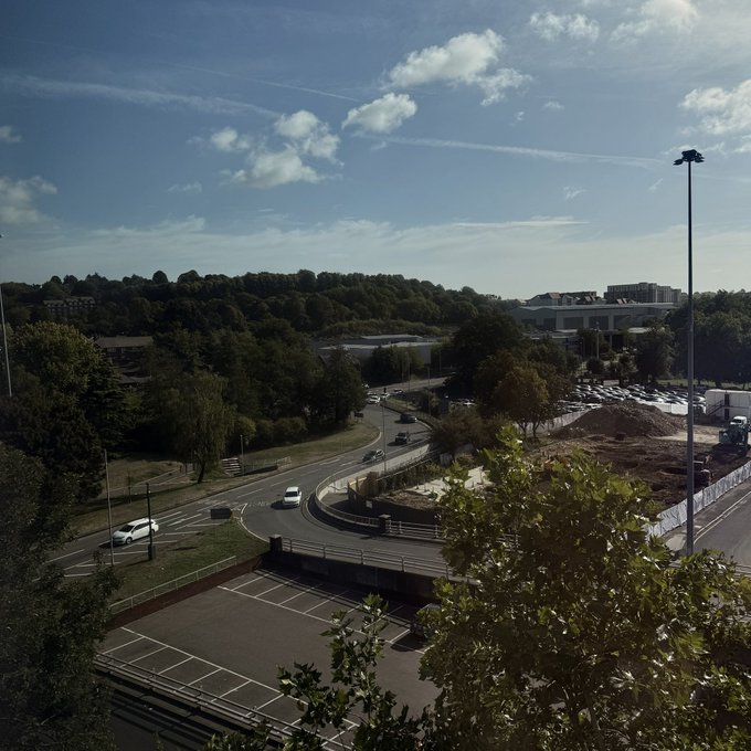 An elevated daytime view overlooking a curving road, parking lot, and wooded hillside under a blue sky with scattered clouds.