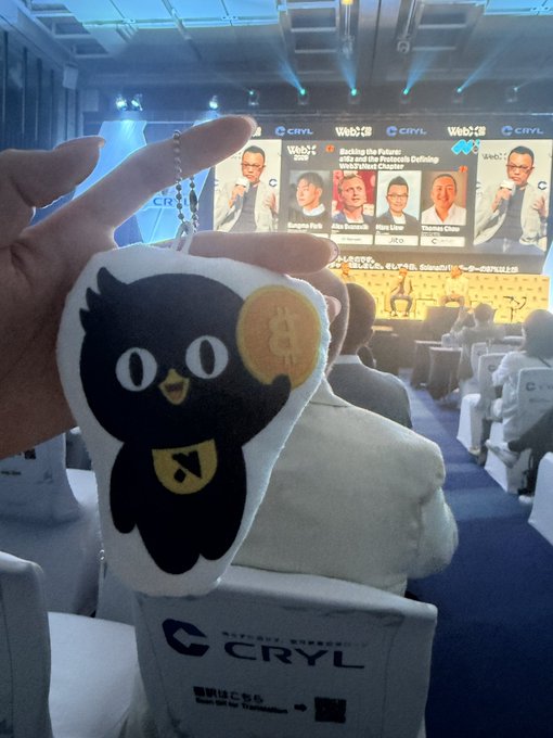 A hand holds up a black cat plush keychain with a Bitcoin coin at a Web3 CRYL conference event, with speakers shown on screens in the background.