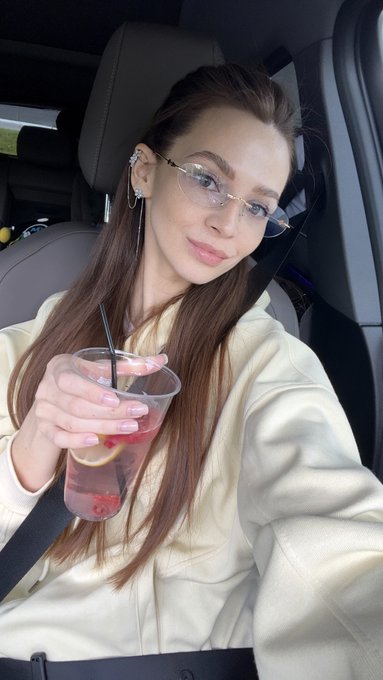 A young woman with long brown hair and glasses sits in a car, holding a clear plastic cup with a pink beverage and a black straw.