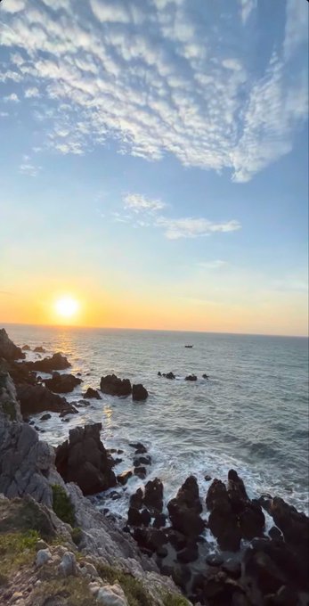 A scenic photograph of a sunset over the ocean with rocky cliffs in the foreground and textured clouds in the sky.