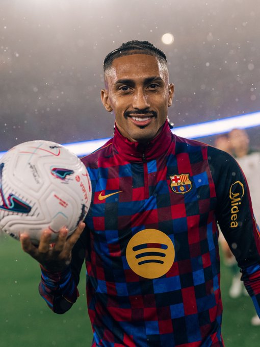 A football player in an FC Barcelona checkered jersey holding a match ball on the field.