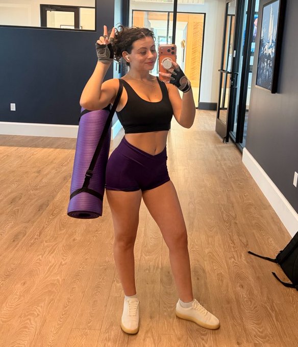 A woman takes a mirror selfie in workout attire (black sports bra and purple shorts) while carrying a purple yoga mat in what appears to be a gym lobby.