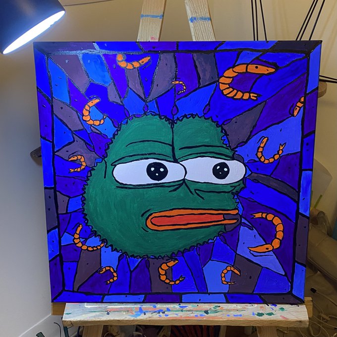 A stained-glass style painting of a green Pepe-like frog face surrounded by a blue and purple shattered-glass pattern with orange worms, displayed on a wooden easel under a studio lamp.