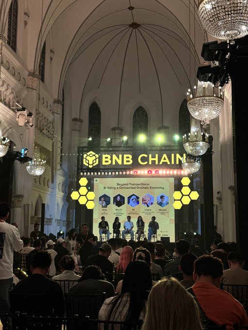 A view from the audience at a BNB CHAIN event titled "Beyond Transactions: Building a Connected Onchain Economy," with panelists seated on stage under ornate chandeliers.