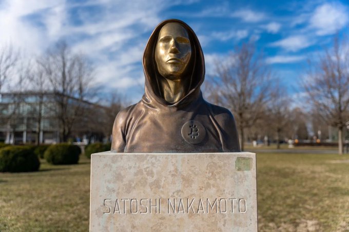 A bronze statue of the hooded figure "SATOSHI NAKAMOTO" stands on a stone pedestal, with a Bitcoin symbol on its chest, embodying the mysterious call to cryptocurrency. The figure's forward gaze and veiled identity visually echo the post's "call beckons me," suggesting an enigmatic invitation into the world of Bitcoin.