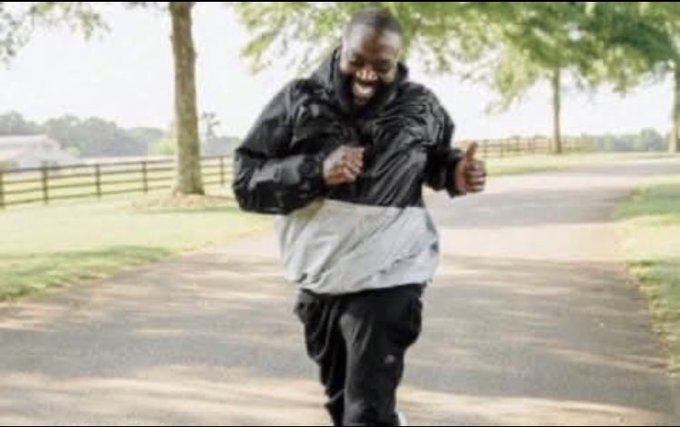 A man in a black and gray jacket jogs along a paved path in a park, giving a thumbs up with a smile.
