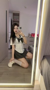 A woman takes a mirror selfie in a bedroom, wearing a white short-sleeved blouse with a black bow, a dark mini skirt, thigh-high stockings, and white sneakers.