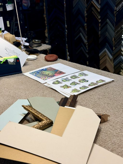 A frame shop workbench displaying custom framing materials, including ornate gold and wood molding samples, mat board, and printed artwork.