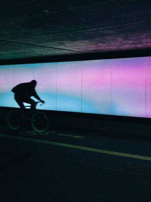 A silhouetted cyclist rides past a wall illuminated with gradient blue, pink, and purple lighting in what appears to be an underpass.
