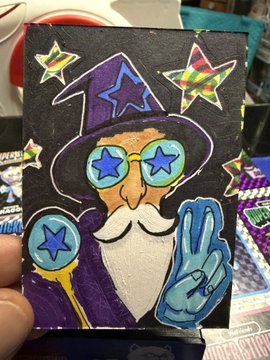 A hand-painted craft or card featuring a wizard character with blue star-shaped glasses, a purple hat, a white beard, and a blue hand making a peace sign against a black background with stars.