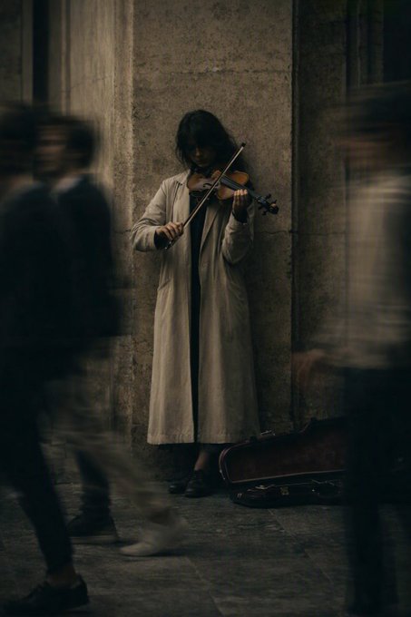 A street musician in a long beige trench coat plays violin against a stone pillar while blurred figures rush past, capturing a quiet, melodic moment amidst urban motion. The image evokes the contemplative "Silent Melody" theme of the new photography collection.