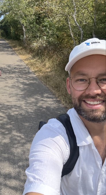 A man wearing glasses, a white cap with the "RWA" logo, and a white shirt with a black backpack strap takes a selfie on a tree-lined road on a sunny day.