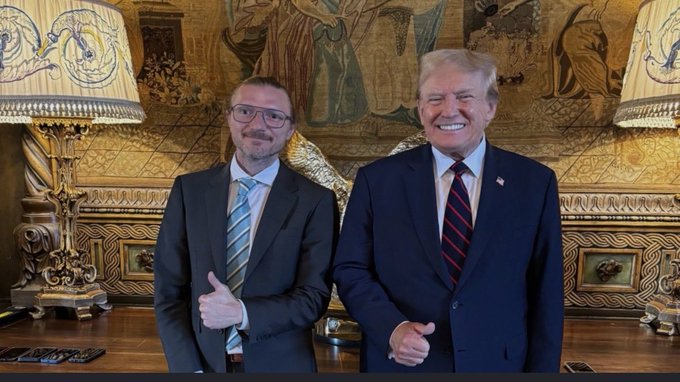 A photo of two men in suits giving thumbs up while standing in an ornate room with tapestry-decorated walls.