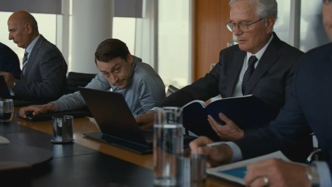 Business professionals in suits working at a conference table, with one man looking at a laptop and an older man reading a document.
