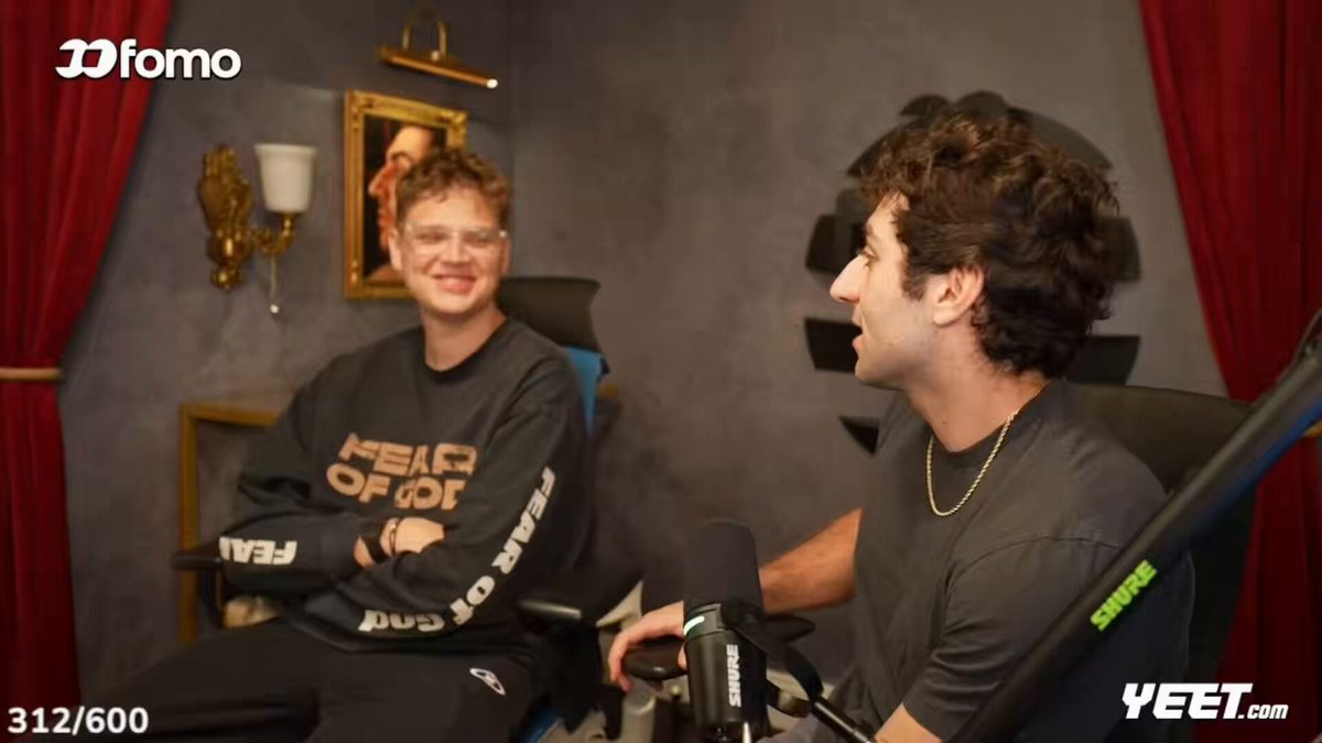 The image shows two men sitting next to each other with microphones in front of them, suggesting they are participating in an interview or discussion. One man appears to be wearing glasses while the other has curly hair. The background features a red curtain and a gold-framed mirror on the wall behind them.