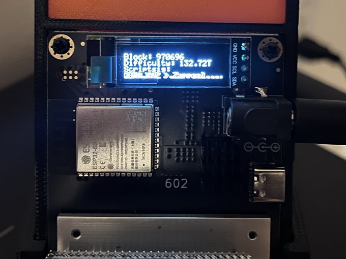 Close-up of an electronic circuit board featuring an OLED display showing text including 'Block: 970696', 'Difficulty: 132.72T', and a worker name, alongside an ESP32-S3-WROOM-1 module.