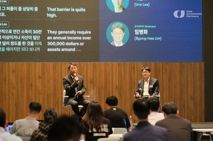 A panel discussion with two men seated on stage in front of an audience, with a large screen behind them displaying translations about 'annual income over 300,000 dollars', and identifying 'Uno Lee' and moderator 'Byung-hwa Lim' alongside 'Kala Investment Partners'.