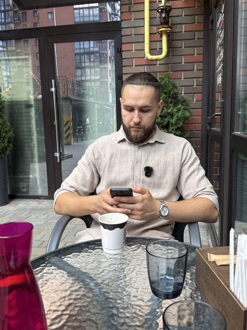 A bearded man sits at an outdoor cafe table looking at his smartphone, with a coffee cup and a glass on the glass table in front of him.