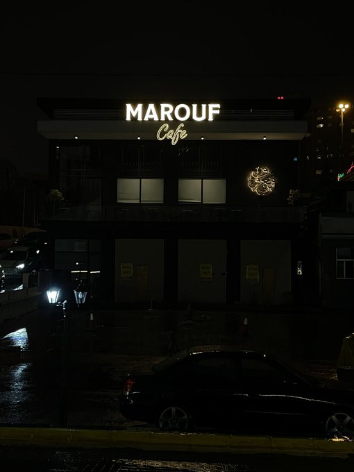 The illuminated storefront sign reads "MAROUF Cafe" in bright white lettering against the night sky, with a dark sedan parked on the wet street below. The image shows no connection to the Rialo/Web3 post, as it simply depicts a nighttime view of a cafe rather than any technology or branding related to that caption.