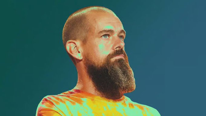 A close-up portrait of a bald man with a full dark beard wearing an orange shirt, lit with teal and orange tones against a teal background.