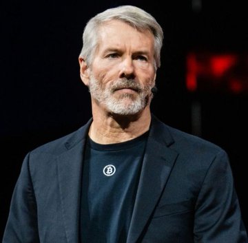 Michael Saylor, the Bitcoin advocate and MicroStrategy executive known for his bullish price predictions, speaks at an event while wearing a dark blazer over a black shirt featuring the Bitcoin logo pin. His appearance aligns with the post's content about his thesis on Bitcoin surpassing gold's market cap by 2035.