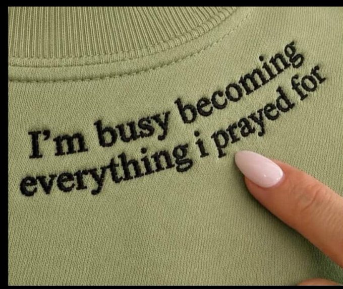 A close-up of an olive green embroidered sweatshirt that reads 'I'm busy becoming everything I prayed for,' with a manicured finger pointing at the text.