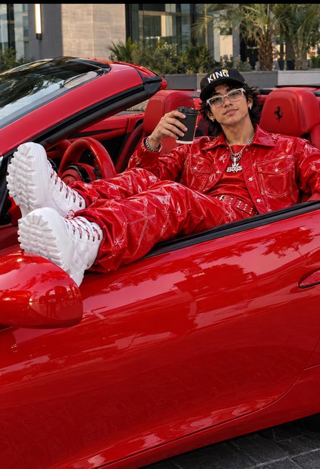 A young man wearing a red vinyl/leather outfit, white sneakers, and a black 'KINR' cap, sitting in a red Ferrari convertible while holding a phone.