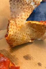 A close-up shot shows a partially eaten slice of pizza crust resting on a cardboard surface.