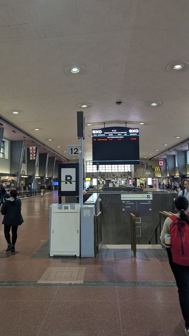 An interior shot of an airport terminal showing a gate area marked with the number 12, featuring a digital flight information display screen above a check-in or boarding counter.