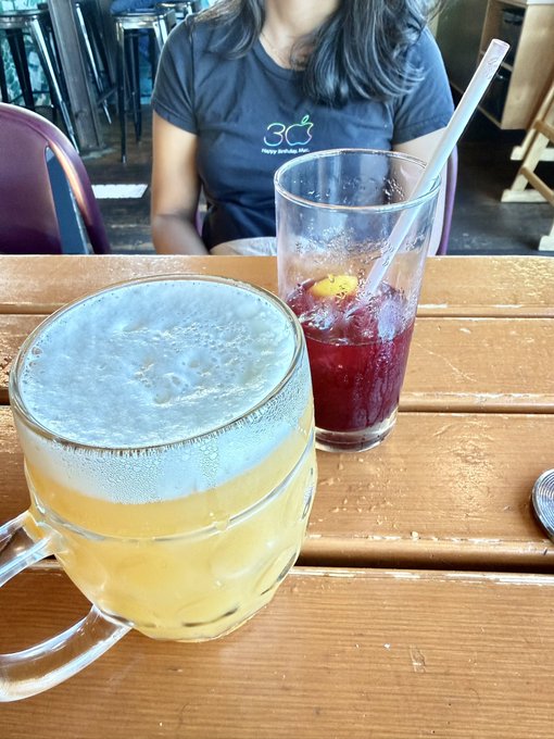 Two refreshing drinks—one a frothy pale ale in a glass mug and the other a vibrant red beverage with a straw and fruit garnish—sit on a wooden table, perfectly embodying a well-earned weekend treat.