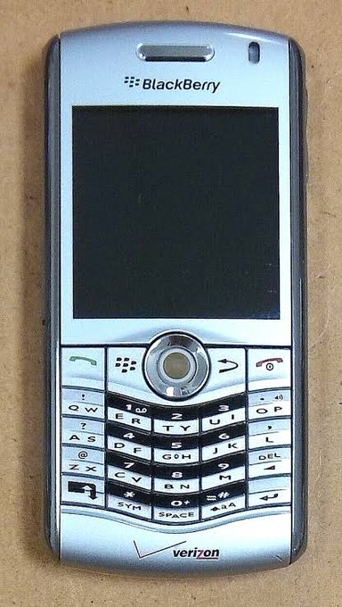 A silver BlackBerry phone, branded with Verizon, rests on a tan surface, its full QWERTY keyboard and "BlackBerry" logo clearly visible, ready to receive the "noods" requested in the post on this classic device.