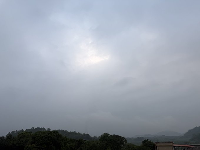 A heavily overcast, gray sky dominates the landscape, with the sun faintly glowing through the thick cloud cover above a silhouette of green hills and trees.