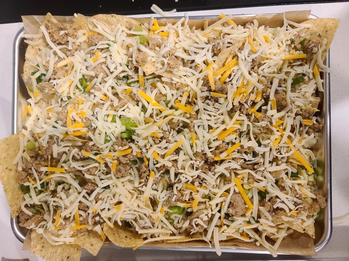 A sheet pan of loaded nachos on a baking tray, featuring tortilla chips topped with shredded chicken, green peppers, and a generous layer of shredded white and yellow cheese, ready to be baked.