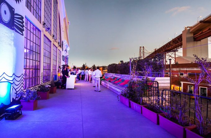 An outdoor rooftop terrace at dusk with purple/blue ambient lighting, decorated with murals, string lights, and plants, with a bridge visible in the background.
