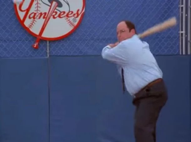 A man in a light blue shirt and tie is shown mid-swing with a wooden baseball bat in front of a blue wall displaying a 'Yankees' logo.