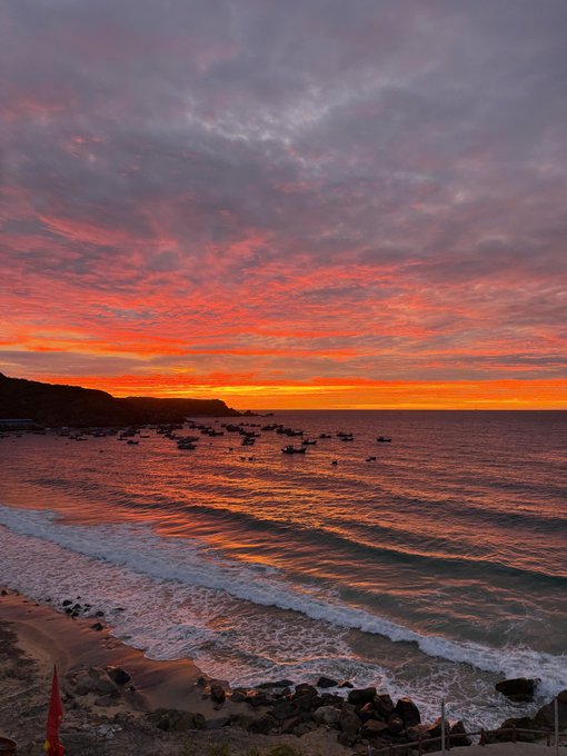 A scenic sunset over the ocean, featuring vibrant orange and purple clouds reflecting on the water with small boats visible on the horizon.