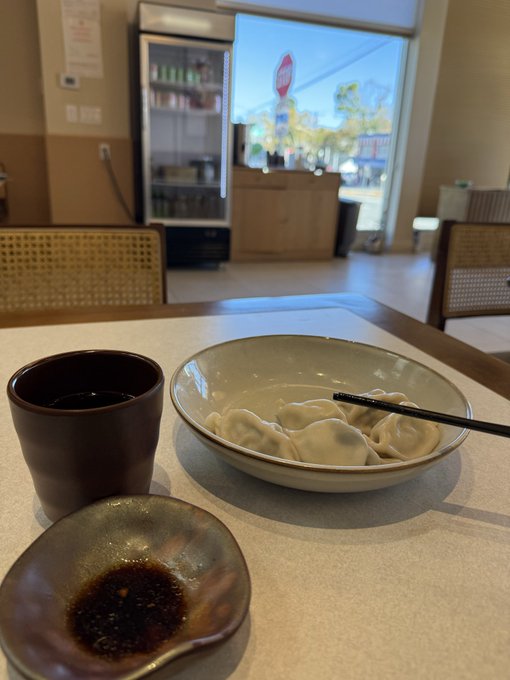 A restaurant table holds dumplings in a bowl, a dark cup, and a small dish of dipping sauce, with a refrigerator and street-facing entrance in the background.