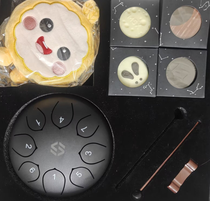 A flat-lay photo of a Mid-Autumn Festival limited edition gift box from MSX/MaiTong, featuring a yellow MaiMai doll cookie with embroidered face, four space-themed chocolate discs with constellation patterns (including a rabbit/bunny and moon designs), a black tongue drum numbered 1–7, and a pair of black mallets with copper-colored handles, all arranged on a dark surface to showcase the starry track packaging.