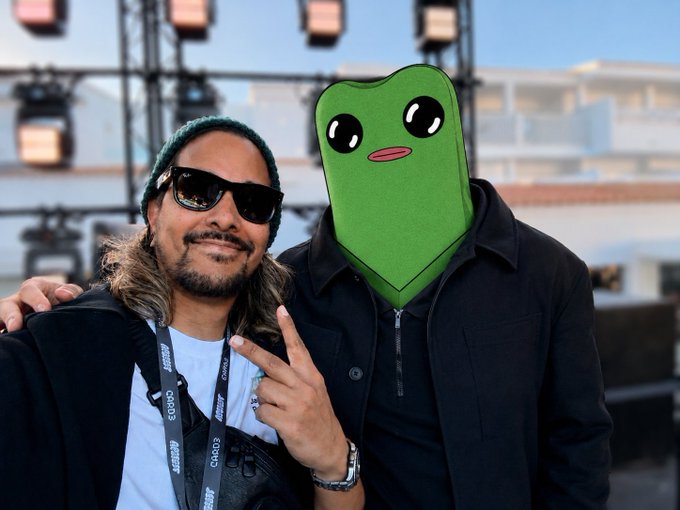 A man wearing sunglasses and a beanie poses with his arm around a person in a green cartoon frog head costume, making a peace sign in front of stage lighting equipment. A "deCODE" lanyard is visible, tying into the crypto-focused "GM fwogs" greeting.