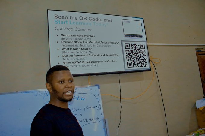 A man stands in front of a TV displaying a slide titled 'Scan the QR Code, and Start Learning today! Our Free Courses:' listing Blockchain Fundamentals, Cardano Blockchain Certified Associate (CBCA), What Is Open Source?, Staking Rewards & Calculation, and Aiken: eUTxO Smart Contracts on Cardano, alongside a QR code.